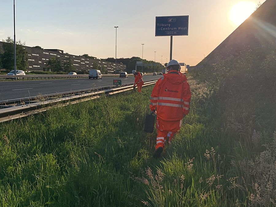 Twee mannen in oranje werkkleding lopen door de berm langs de snelweg