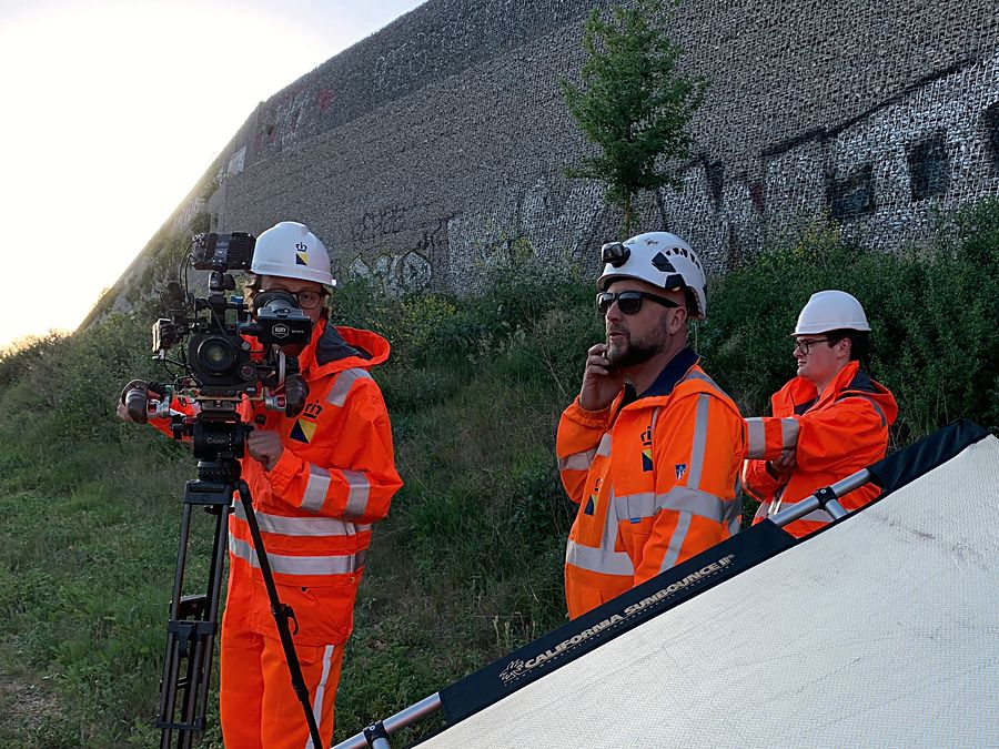 Cameraman op statief langs het spoortalud, ploeg in oranje werkkleding erbij